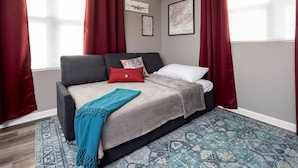 A bedroom with a grey sofa bed, red and white pillows, a blue throw blanket, and a patterned rug. There are two framed pictures on the wall and a window with red curtains. - Modern City Oasis: Pet Friendly | Downtown | Park (Little Rock)