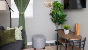 Een gezellige zithoek met een grijze bank, een klein rond tafeltje en een grijze poef. Er hangt een groen gordijn, staat een grote groene plant en is een ronde wandklok.