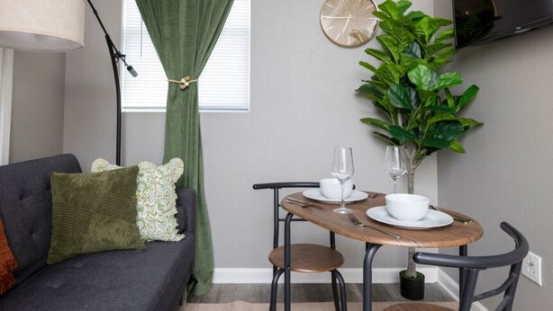 Een kleine woonkamer met een grijze bank, een houten tafel met twee stoelen, een kleine potplant en een aan de muur bevestigde televisie.