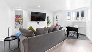 Un salón moderno con un sofá gris, un televisor de pantalla plana, una pintura sobre caballete y una mesa de comedor con sillas.