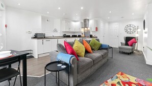 Una sala moderna con un sofá gris, cojines coloridos, una mesa de comedor con sillas, una mesita auxiliar, una zona de cocina con armarios blancos, un reloj y una lámpara.