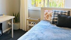 A bedroom with a bed, a desk, a potted plant, and a window with a view of a bamboo fence.