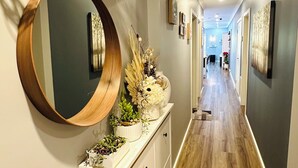 A hallway with a wooden floor, a white console with decorative plants, a round mirror, and a red vase.