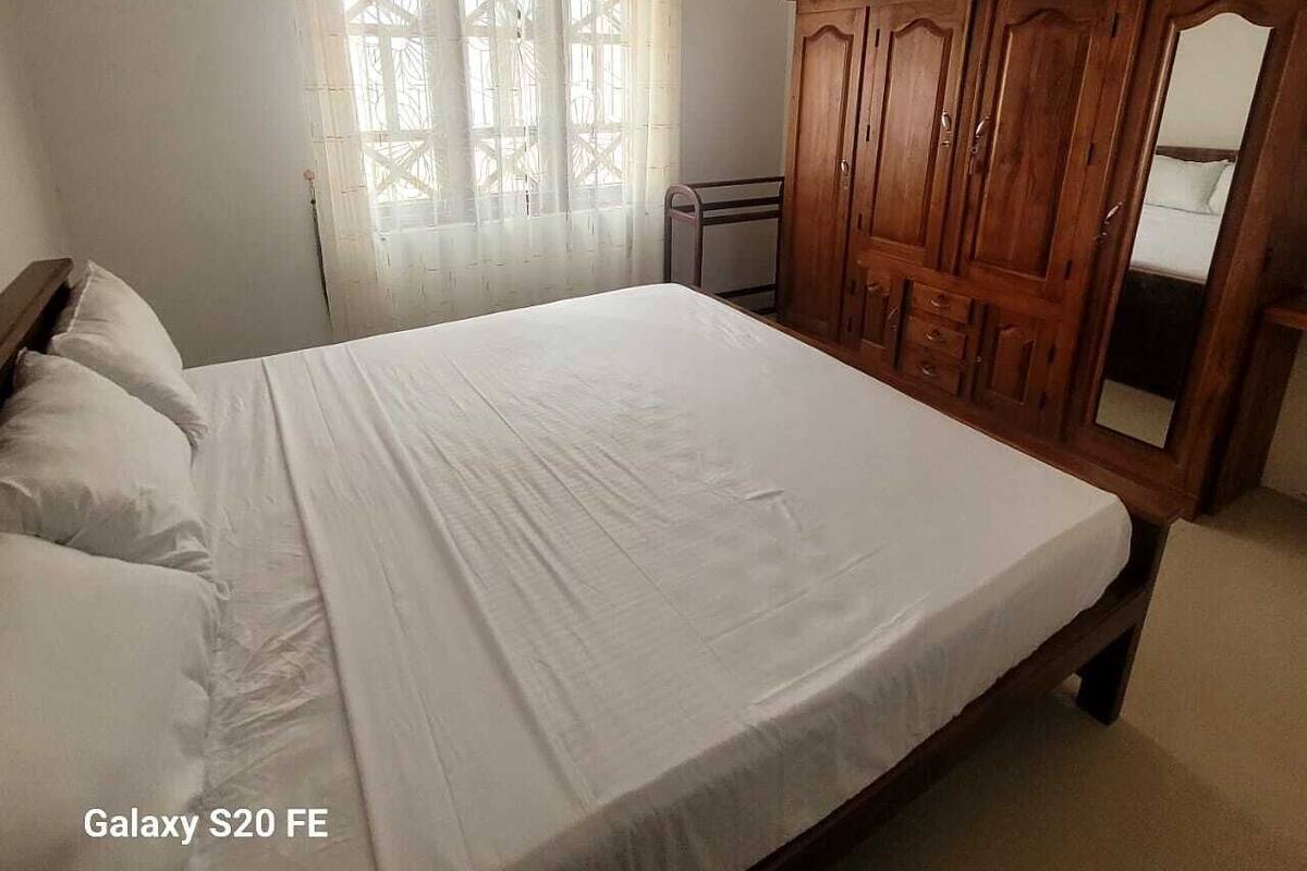 A bedroom with a Galaxy S20 FE bed, a wooden wardrobe, and a window with sheer curtains.