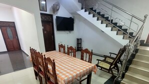 A dining area with a checkered tablecloth, wooden chairs, a staircase with metal railings, and a wall-mounted TV. - Grace- The Joyful Rest (Batticaloa)