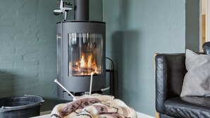 Een woonkamer met een zwart houthakkerkachel, een zwarte leren bank en een witte tafel met een plaid en tijdschriften.