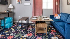 Un accogliente soggiorno con un divano blu, un tavolino in legno apparecchiato con piatti e bicchieri, un tappeto floreale e un piccolo tavolino laterale con una lampada.