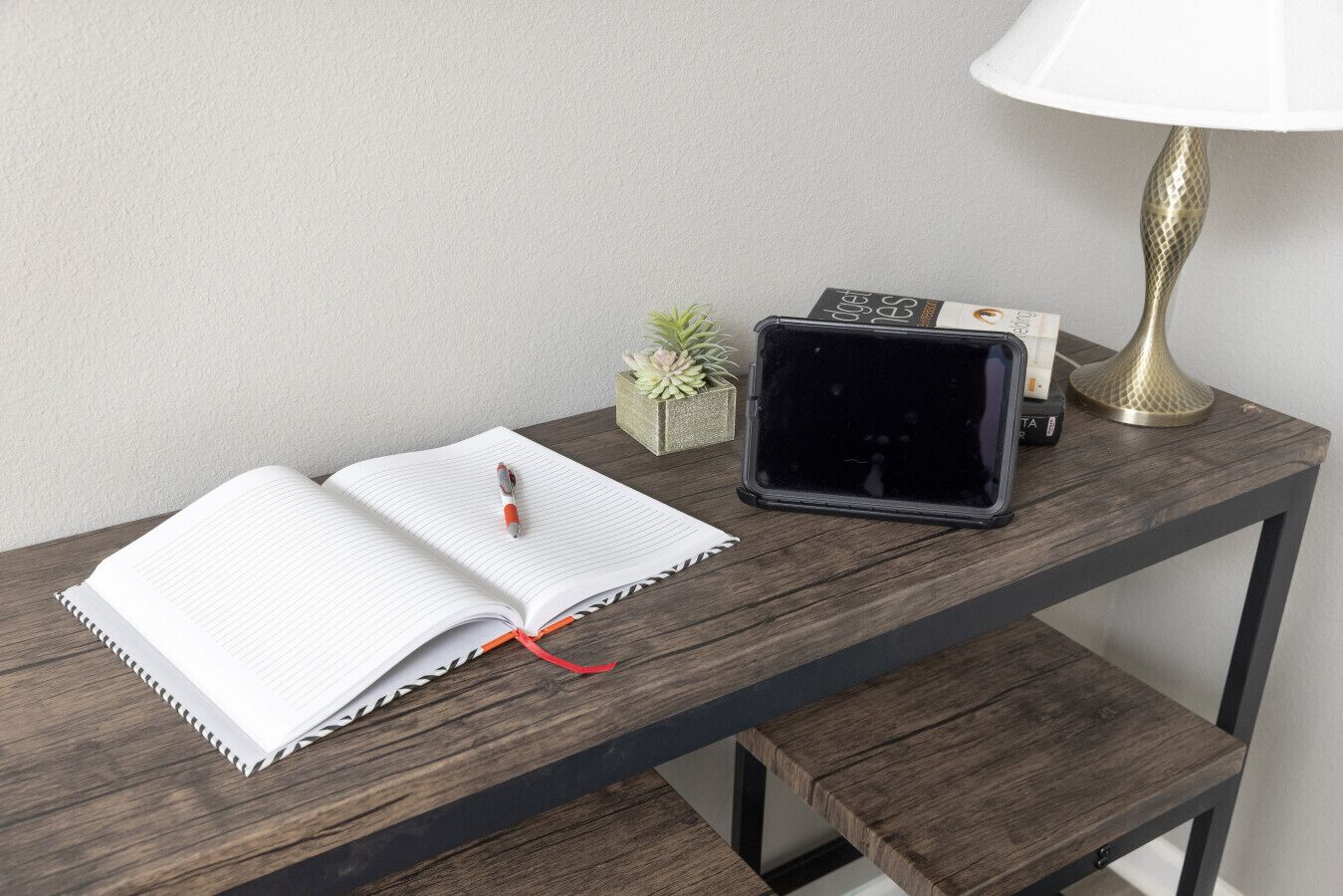 A wooden desk with a notebook, a pen, a tablet, a lamp, and a small plant.