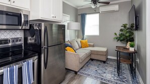 A compact kitchen with a refrigerator, stove, and microwave. There's a beige sofa with cushions, a small table with a plant, and a patterned rug.