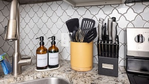 A kitchen with a stainless steel faucet, two bottles of dish soap, a yellow utensil holder with black spatulas and whisks, and a black knife block.