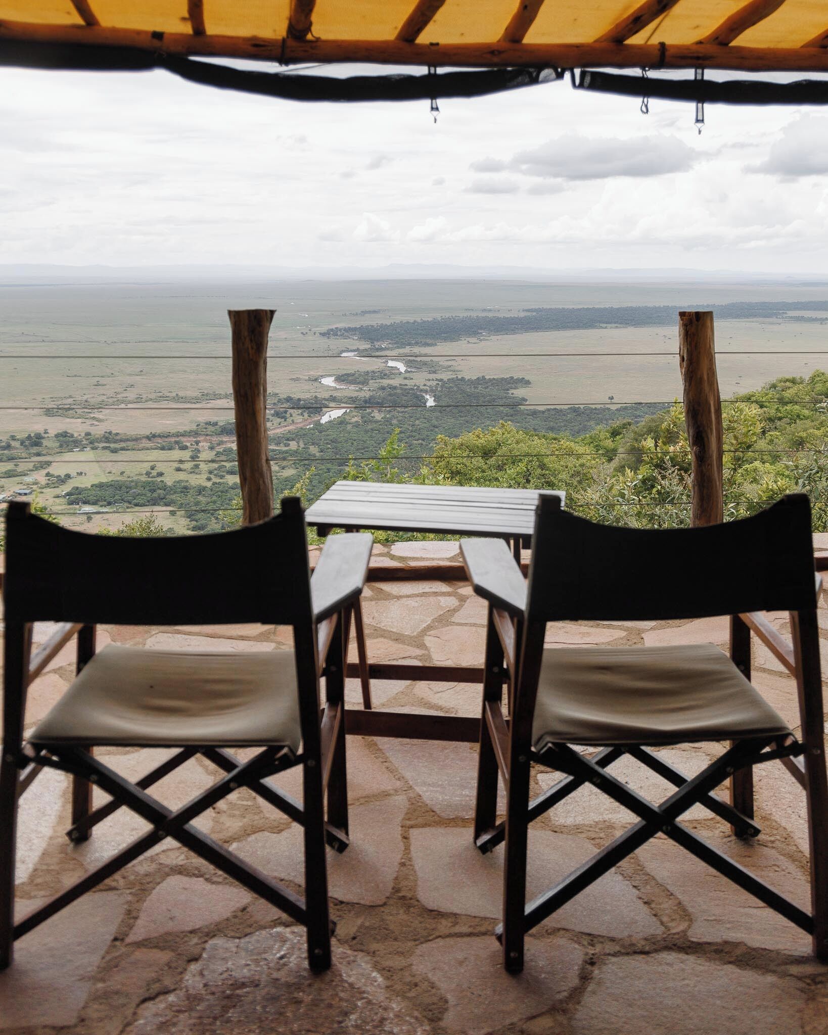 Two chairs and a table under a wooden structure with a view of a river and distant landscape.