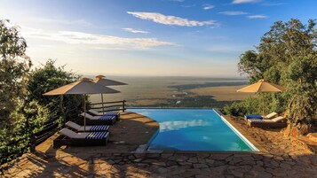 Una piscina con sillones y sombrillas, rodeada de suelo de piedra y árboles, con vistas a un paisaje extenso.