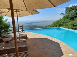 Una piscina con vista al océano y colinas, rodeada de sillas de descanso y una pérgola.