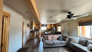 A spacious living area with a large sofa, a coffee table, and a dining table. There is a kitchen area in the background with wooden cabinets and a refrigerator.