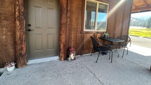 A wooden cabin with a green door, a window, a table with chairs, and a small lantern on the porch.