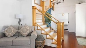 A staircase with wooden railings and a chandelier, a grey sofa with patterned cushions, and a small round wicker basket.