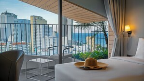 A hotel room with a balcony, a bed, a chair, and a view of the city and ocean. - Seaside Boutique Hotel Nha Trang Beach (Nha Trang)