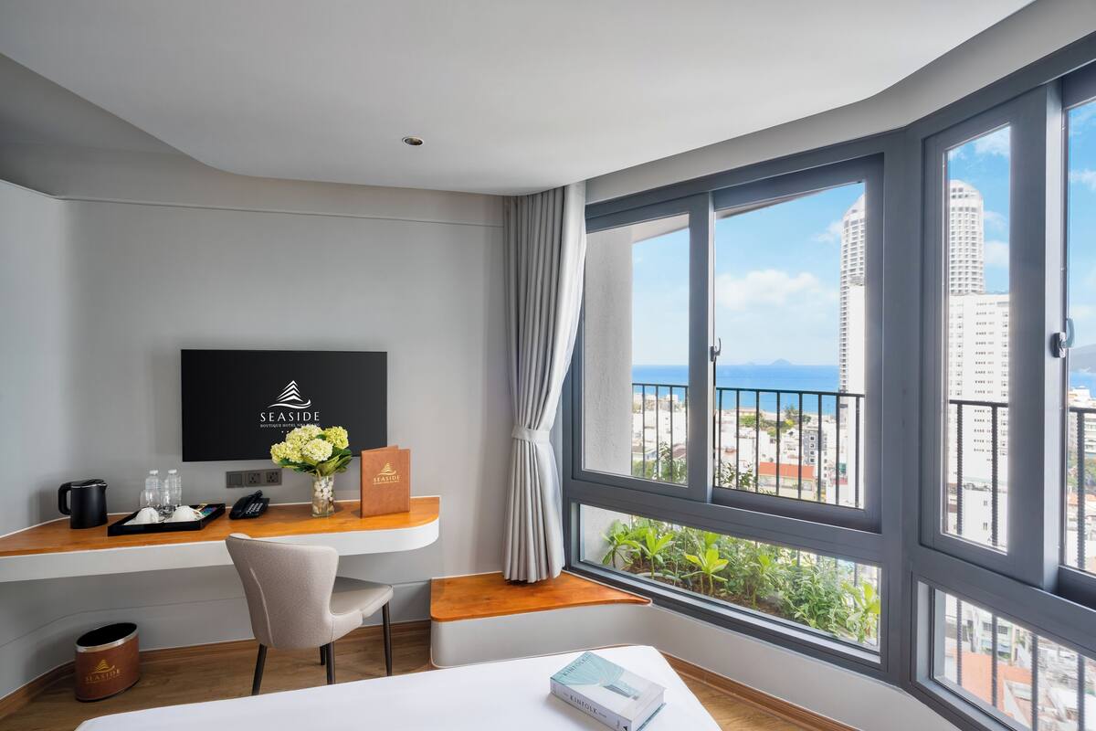 A hotel room with a large window, a desk with a flat-screen TV, a vase with flowers, and a view of the city and sea.