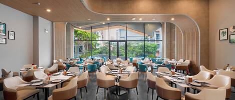 A modern dining area with multiple tables and chairs, large windows offering a view of greenery outside.