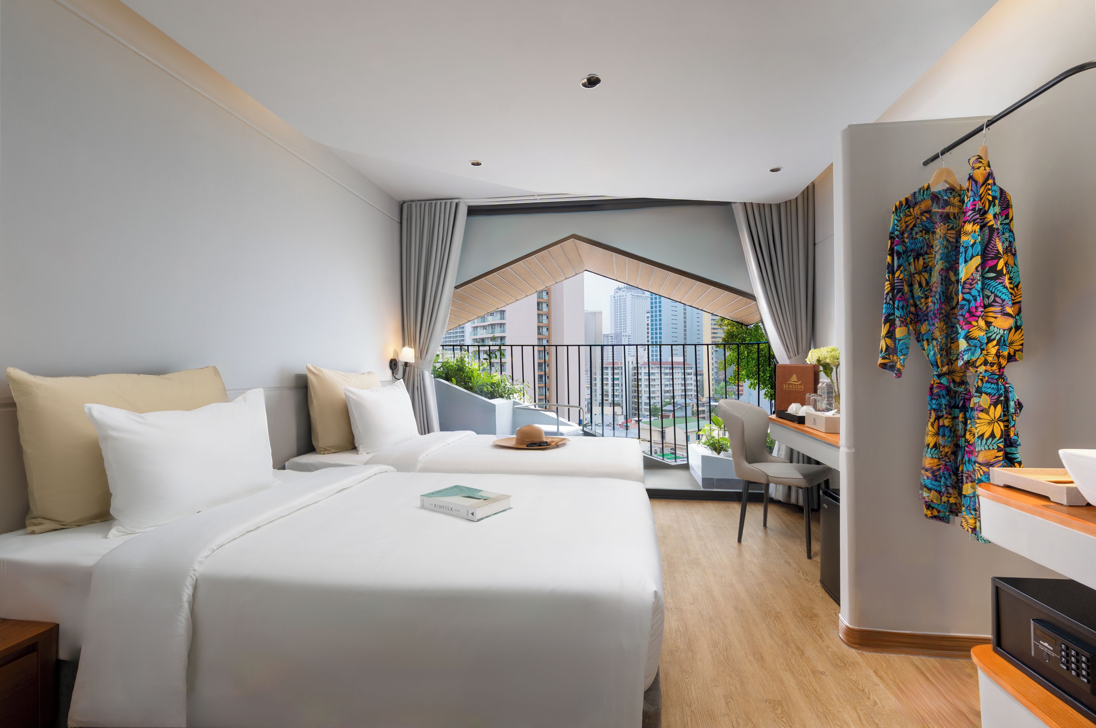 Habitación de hotel con dos camas, un escritorio y vistas a un balcón con plantas y edificios.