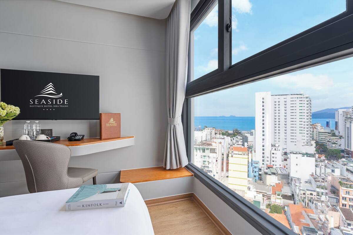 A hotel room with a large window, a desk with a chair, a flat-screen TV, and a book on the bed.