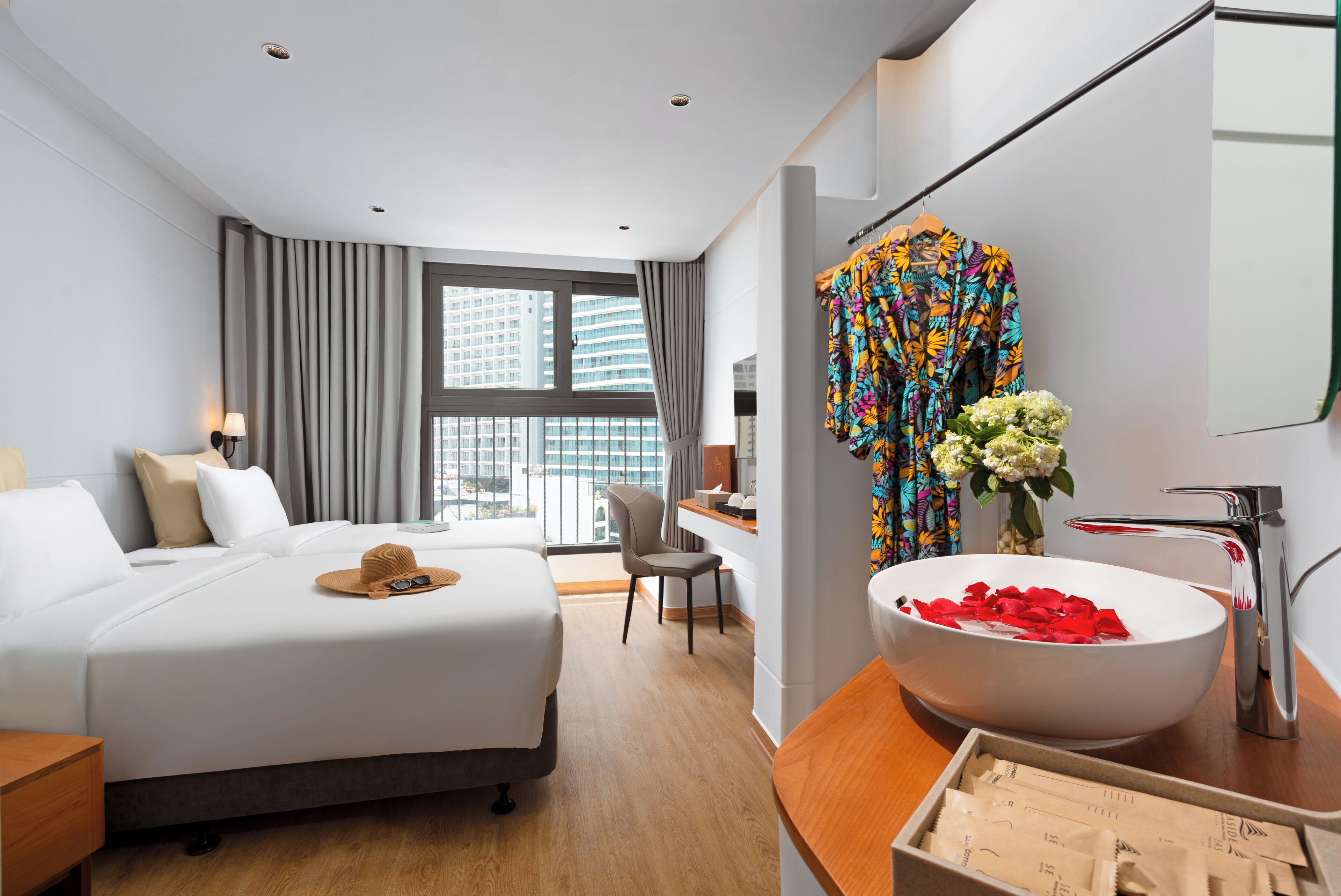 Een moderne hotelkamer met een groot bed, een bureau, een badkamer met een witte wastafel en uitzicht op een stadsbeeld door grote ramen.