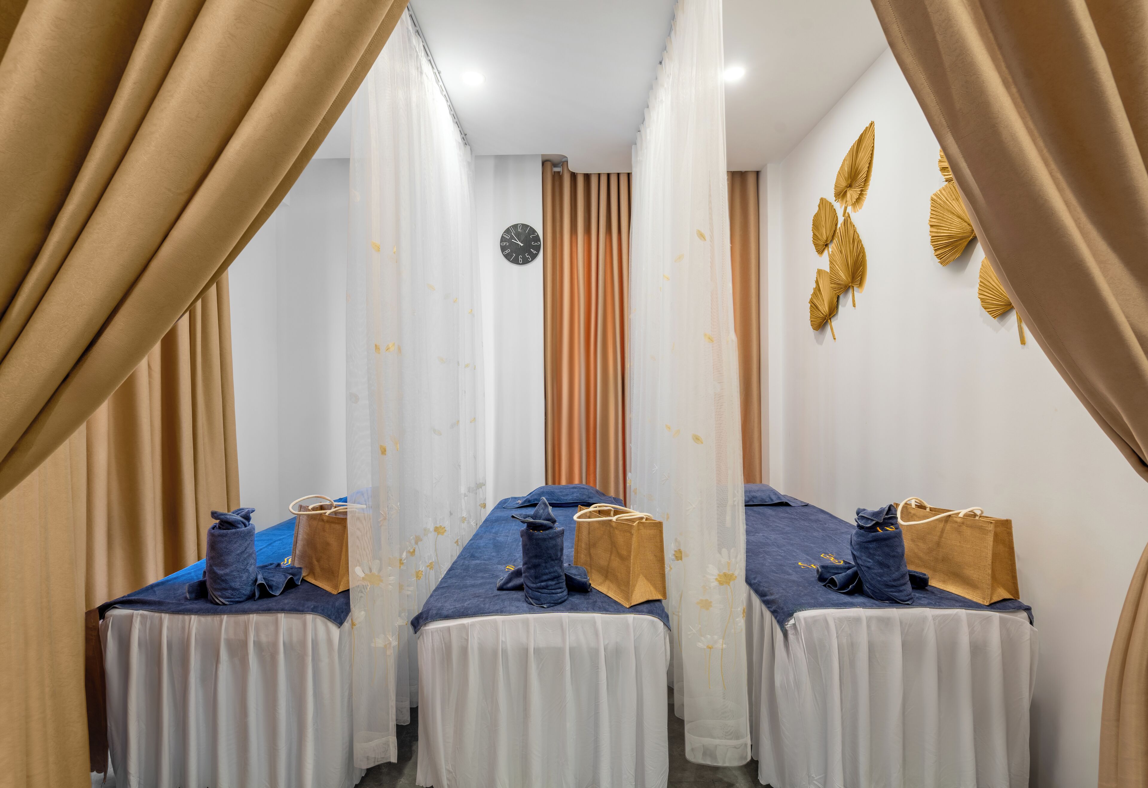 A room with three beds, each with a blue bedspread and white curtains. There are brown bags on the beds, and a clock on the wall.