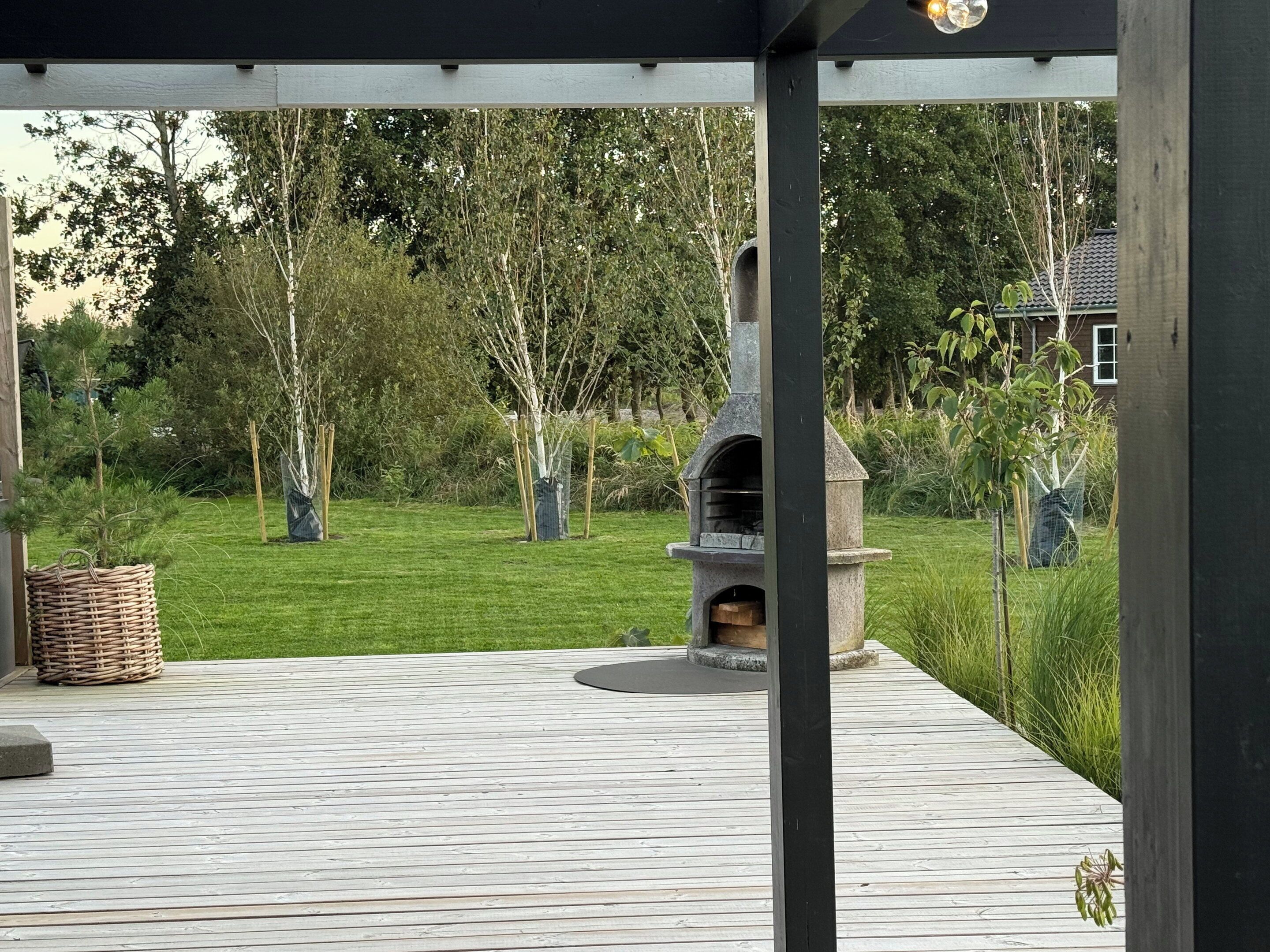 Ett takförsett utomhusområde med en grillplats av sten, ett trädäck och en gräsyta.