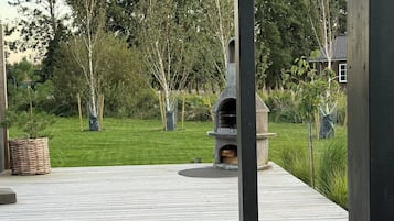Un espace extérieur abrité comprenant un barbecue en pierre, une terrasse en bois et une pelouse.