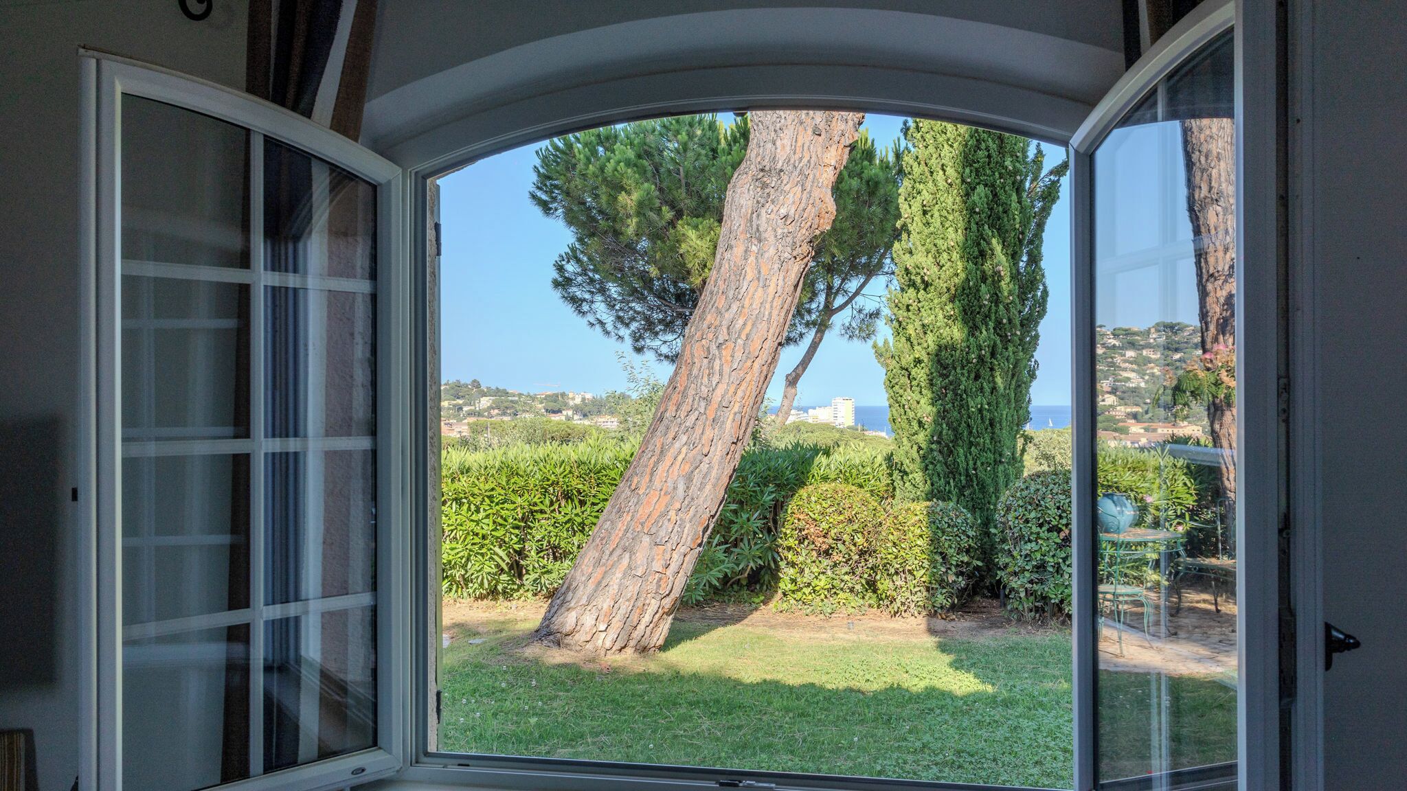 Une pièce dotée d’une grande fenêtre en arc d’où l’on peut admirer un jardin avec un grand arbre, une haie et une côte lointaine.