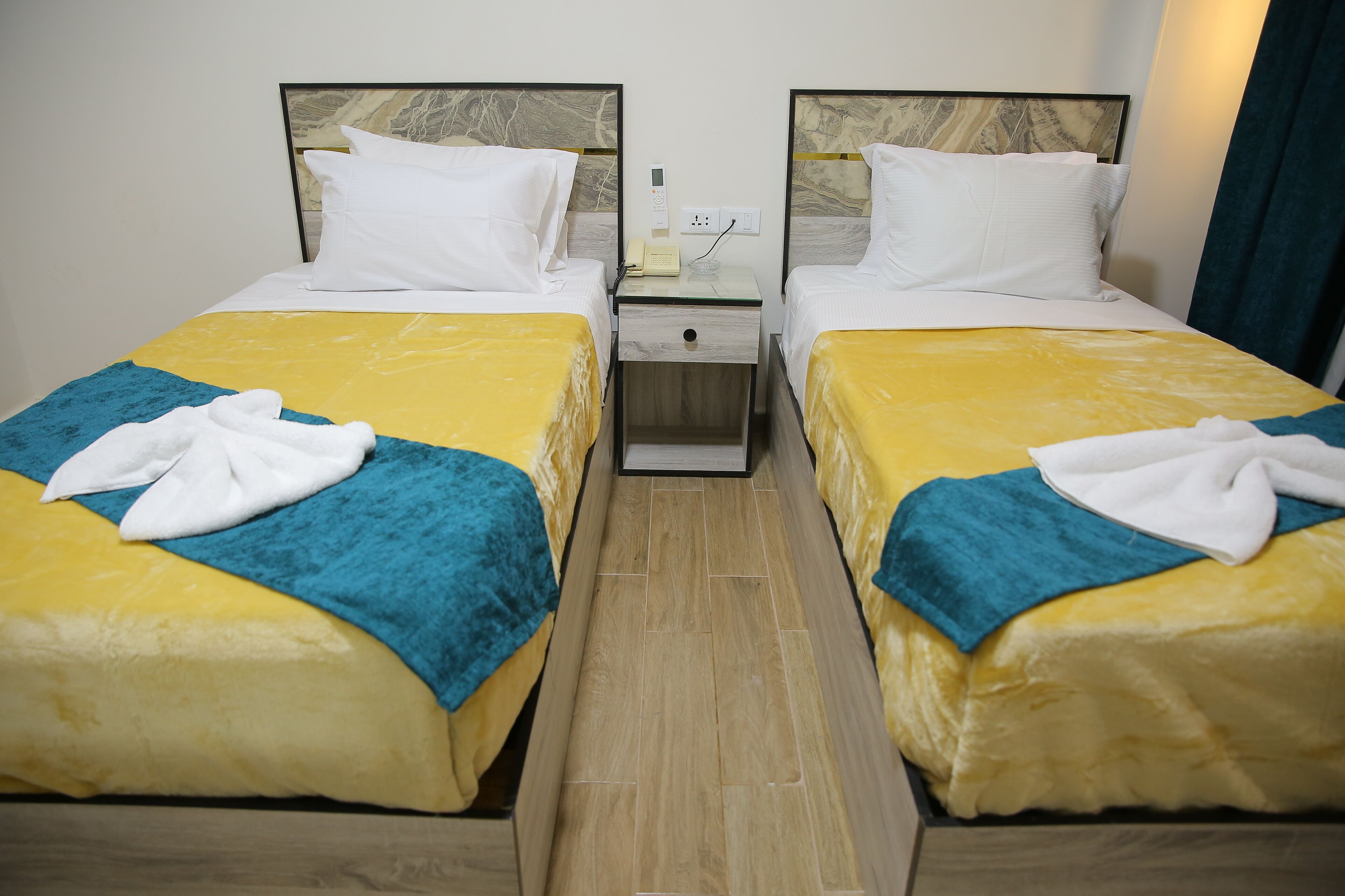 A hotel room with two beds, each with a yellow and blue bedspread, white pillows, and a white bathrobe on the bed. There is a nightstand with a phone and a power outlet between the beds.
