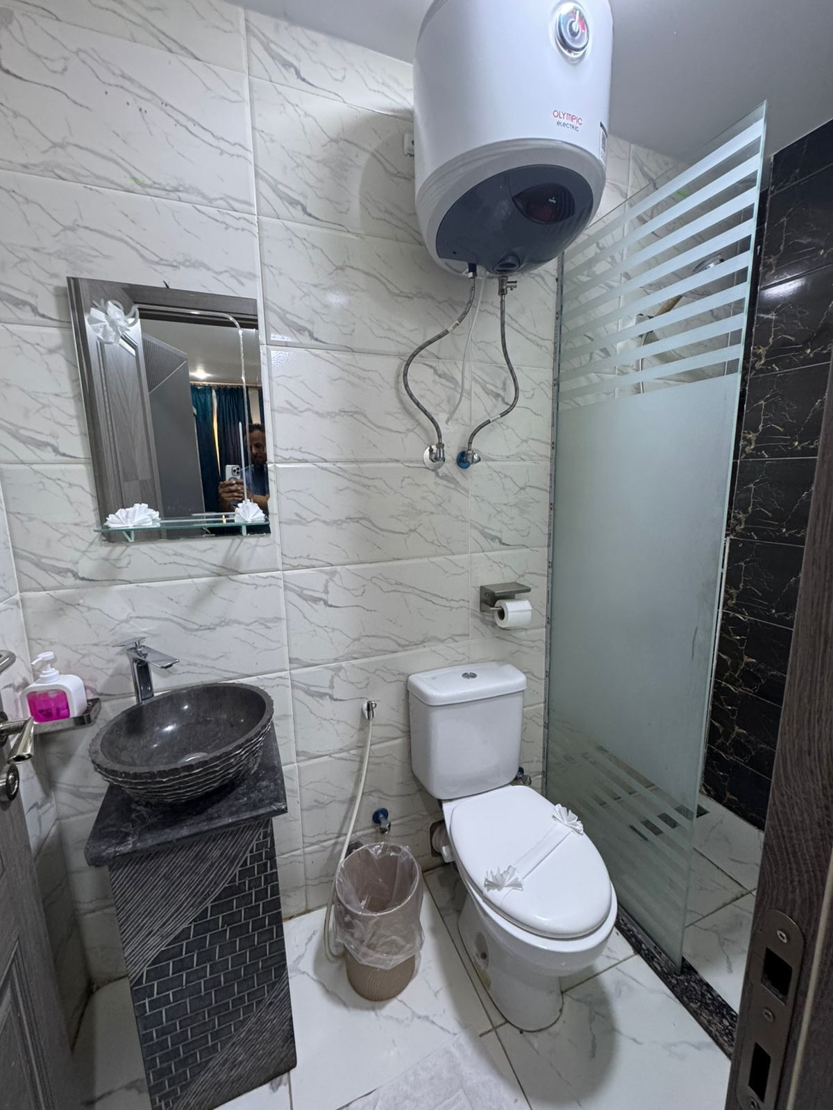 A bathroom with a marble wall, a sink with a basin, a toilet, a shower enclosure, and a mirror with a decorative frame.