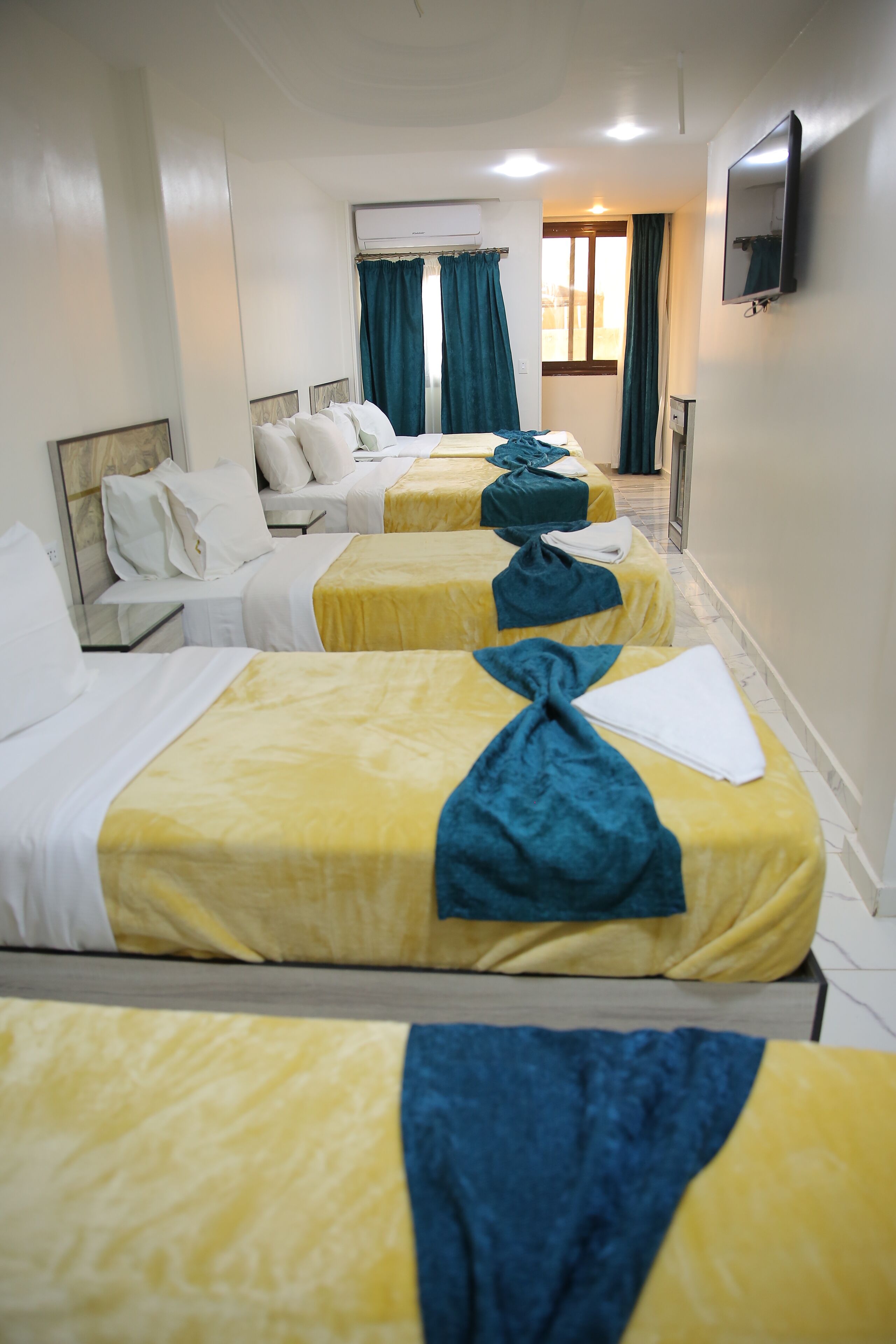 A hotel room with three beds, each with a yellow and blue bedspread, white pillows, and a neatly folded white towel.