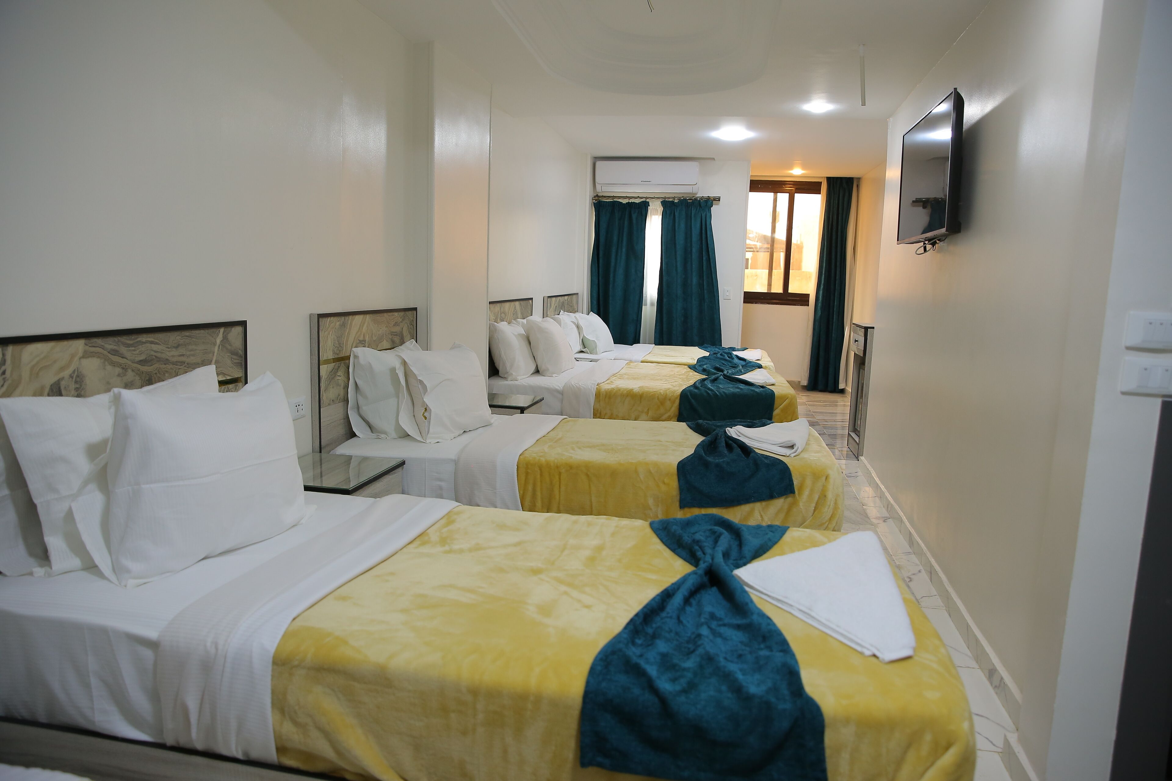 A hotel room with three beds, each with a different colored bedspread, a flat-screen TV mounted on the wall, and an air conditioning unit above the beds.