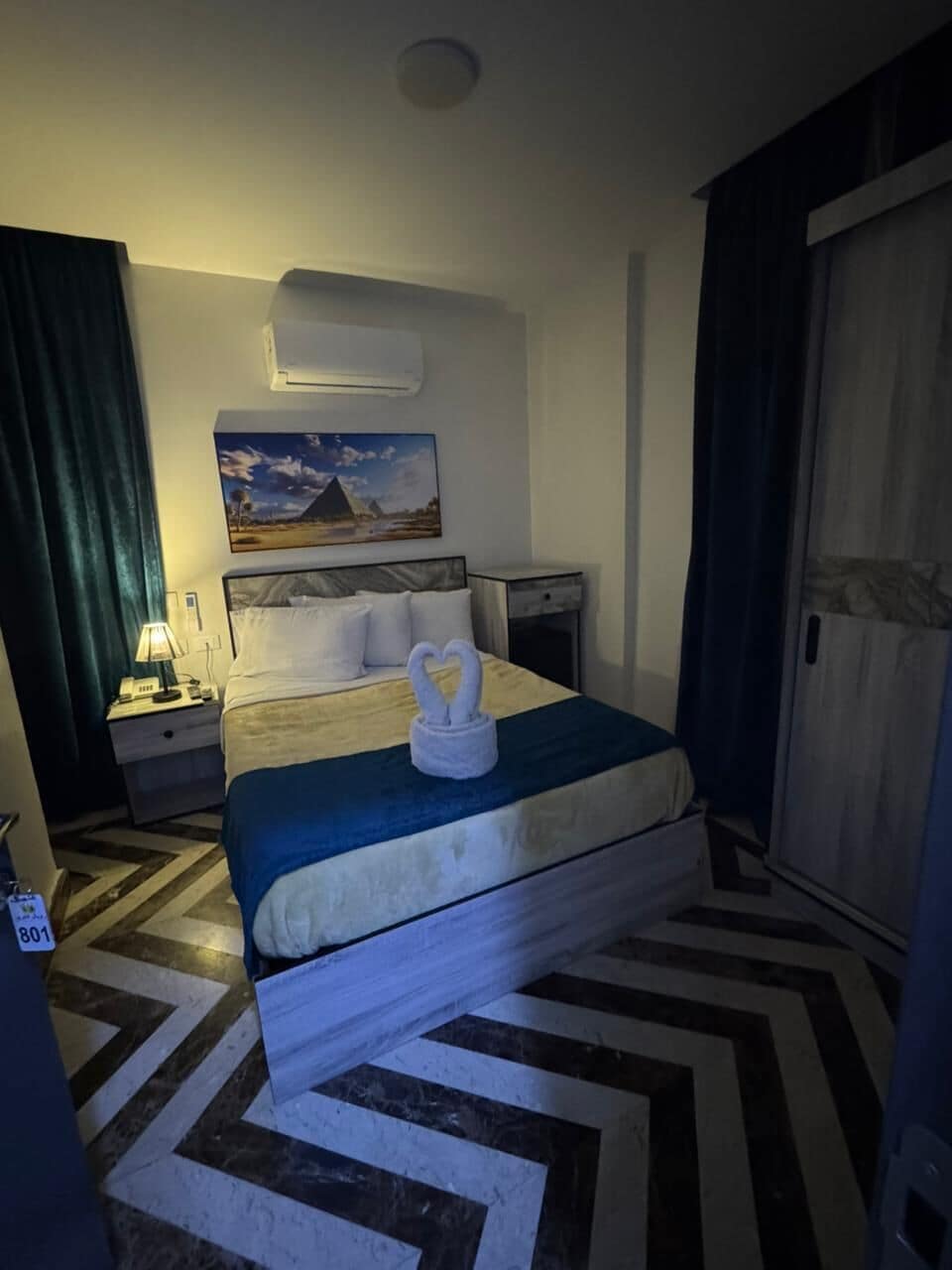 A hotel room with a bed, a nightstand, a lamp, a painting of a pyramid, and a door.