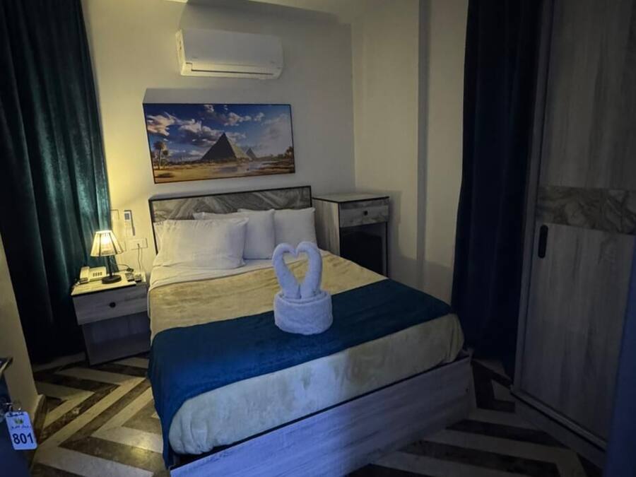 Une chambre d’hôtel avec un lit, une table de chevet, une lampe, un tableau représentant une pyramide et une porte.