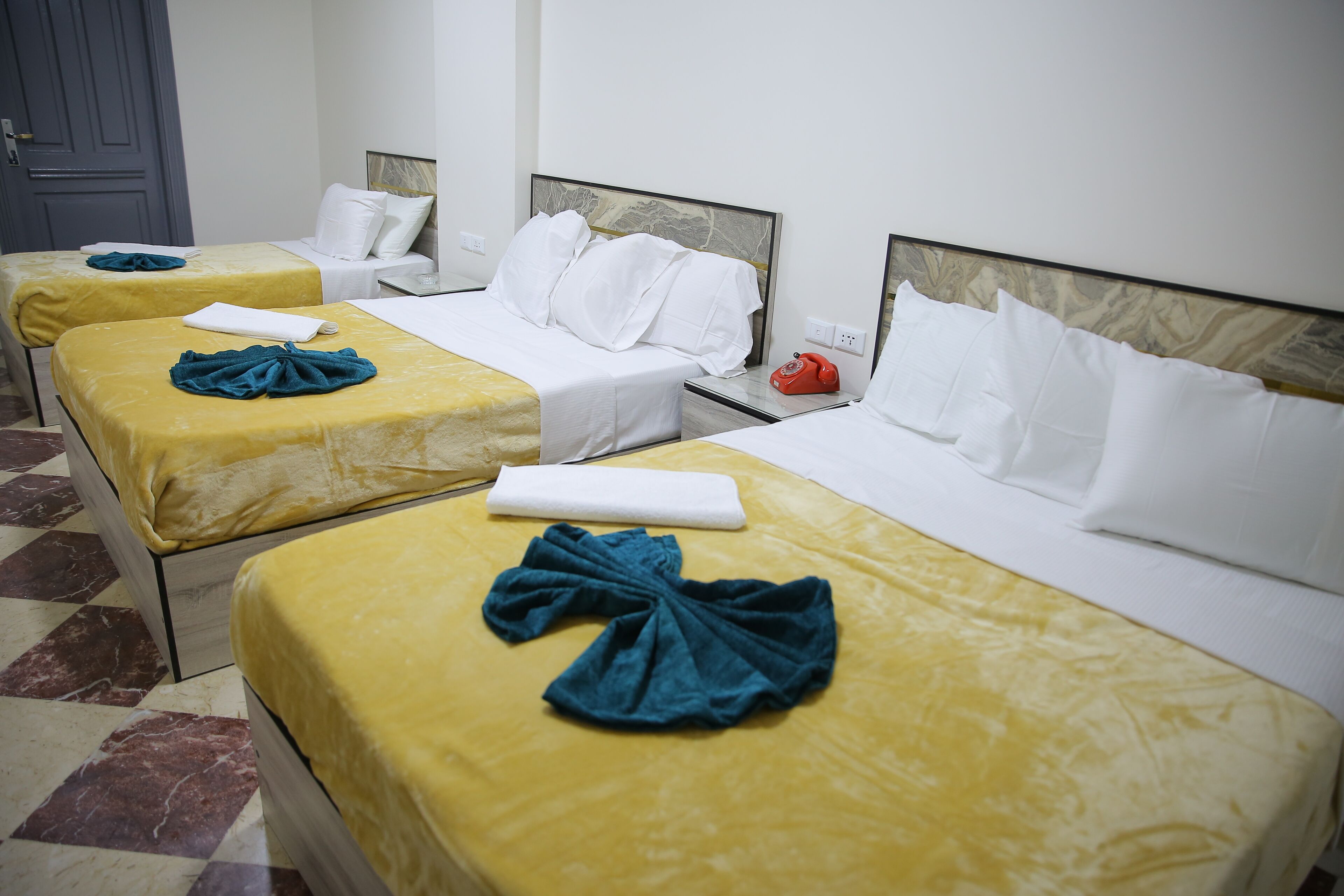 A hotel room with three beds, each with a yellow bedspread and white pillows. There is a telephone on the nightstand and a door in the background.