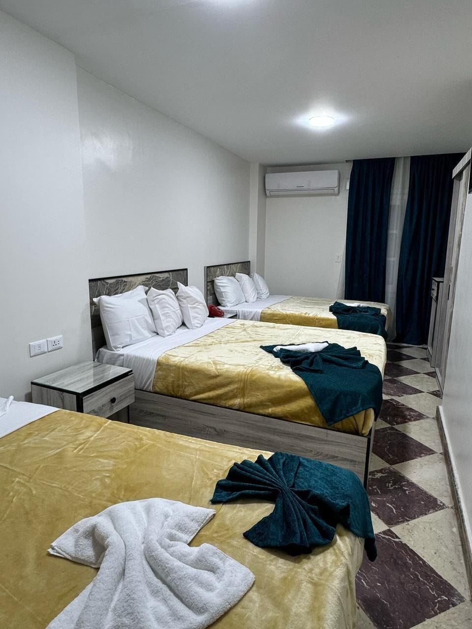 A hotel room with two beds, a bedside table, a wardrobe, and an air conditioning unit.