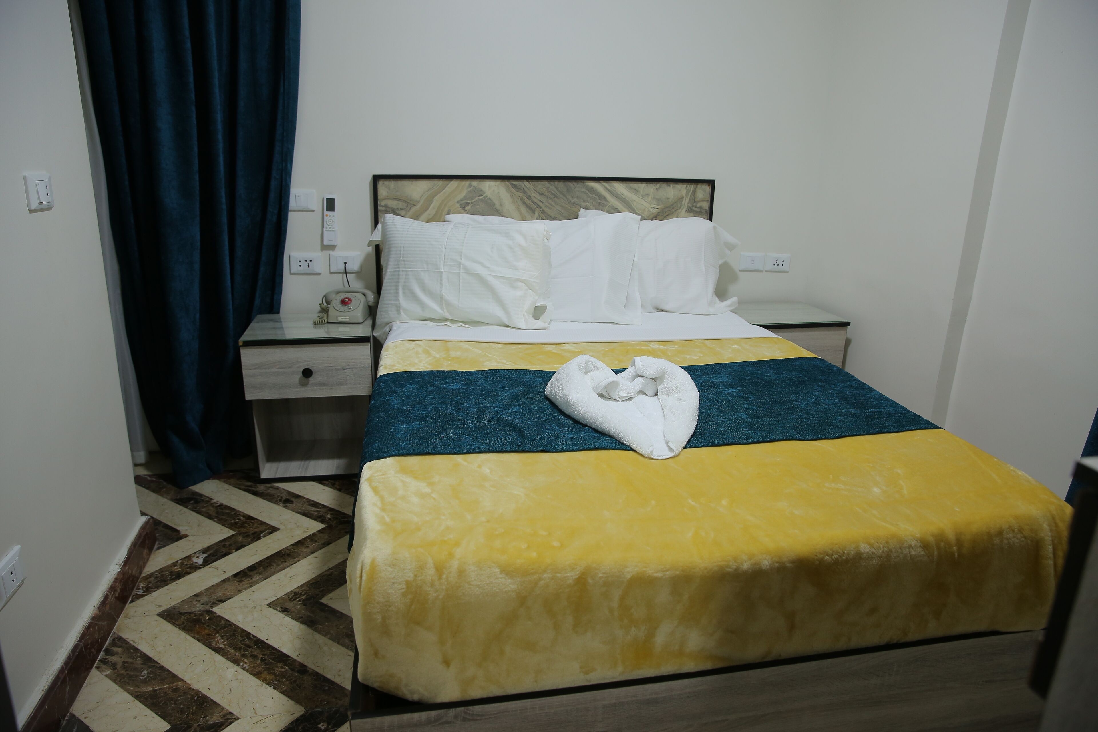 Una habitación de hotel con cama, mesita de noche, lámpara, toma de corriente y cortina azul.