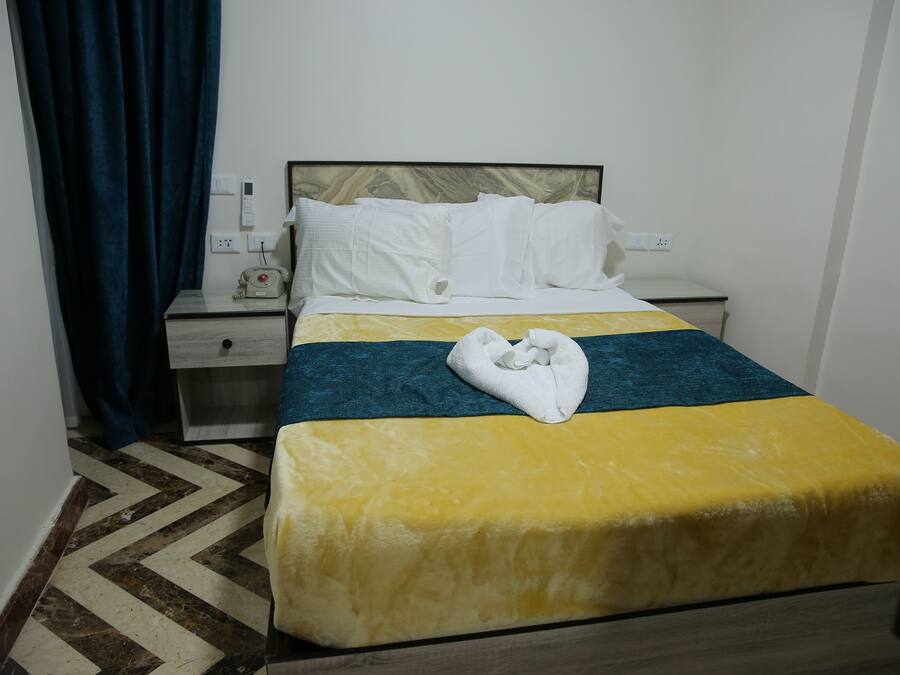 Una habitación de hotel con cama, mesita de noche, lámpara, toma de corriente y cortina azul.