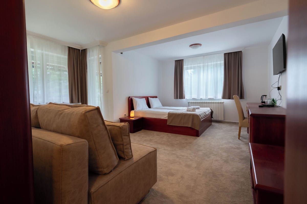 A hotel room with a sofa, a bed with a wooden headboard, a desk, and a television mounted on the wall.