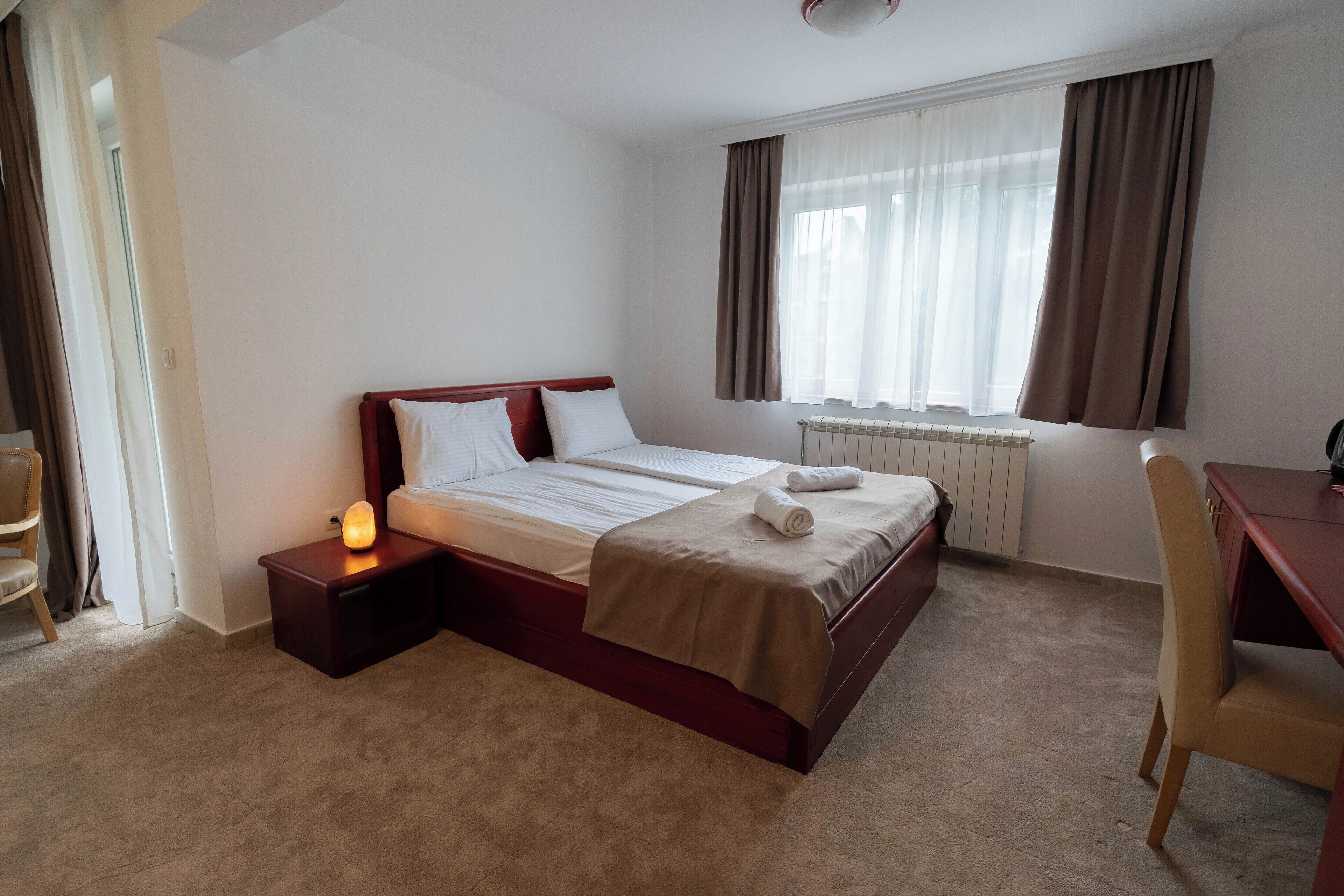 A hotel room with a single bed, bedside table with a lamp, a chair, and a desk.