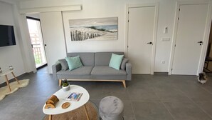 En moderne stue med en grå sofa, et rundt sofabord og en lille, rund skammel. Et maleri af en strandscene hænger på væggen, og der er en dør til højre.