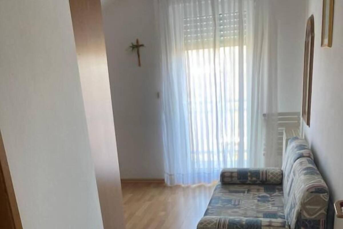 A room with a wooden floor, a sofa with patterned cushions, a cross on the wall, and a window with sheer curtains.