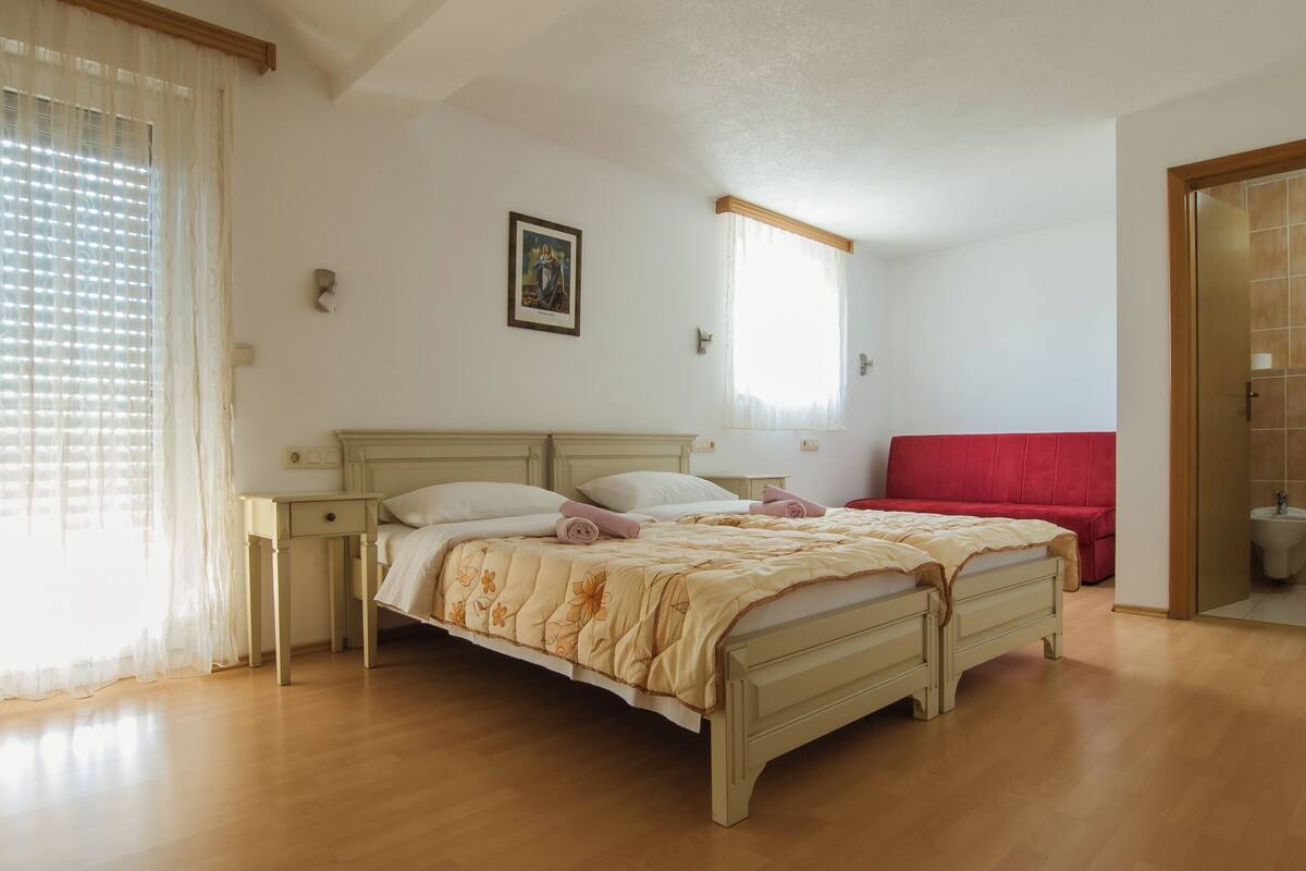 A bedroom with a bed, a nightstand, a red chair, a window with curtains, and a wall-mounted picture.
