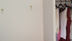 A room with a wardrobe containing hanging clothes, a small wooden chest with various items, and a desk with a keyboard and other objects.