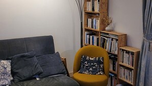 En hyggelig stue med en mørkegrøn sofa, en gul lænestol, et træbord med bøger, en gulvlampe og en reol fyldt med bøger.