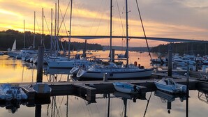 En lystbådehavn med fortøjede både, en bro i baggrunden og roligt vand der spejler himlen.