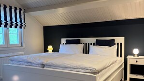 Ein Schlafzimmer mit einem weißen Bett, einer schwarz-weißen Bettdecke, einem Nachttisch mit Lampe und einem Fenster mit Jalousien.