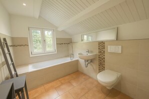 Ein Badezimmer mit Badewanne, Waschbecken und Toilette. Ein Fenster sorgt für natürliches Licht und über dem Waschbecken befindet sich ein Spiegel.